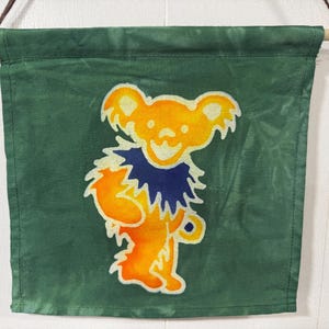 Puede incluir: Bandera verde con un oso sonriente en naranja, amarillo y azul. El oso tiene un cuello azul y está centrado en la bandera. La bandera está colgada de un palillo de madera con una cuerda marrón.