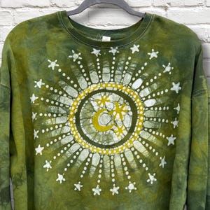 May include: Green tie-dye sweatshirt with a celestial design. The design features a central circle with a crescent moon and star motif, surrounded by radiating lines and star accents. The sweatshirt is hanging on a black hanger.