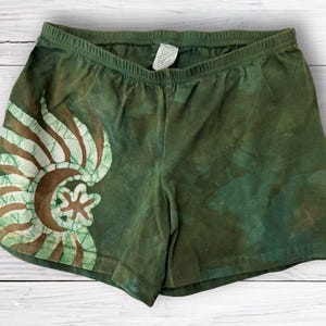 May include: Green shorts with an elasticated waistband and a tie-dye effect. The shorts feature a large, abstract design on the left leg in shades of brown, white, and green. The design resembles a stylized feather or wing.