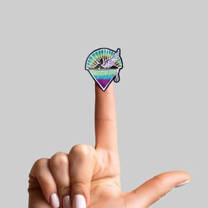 May include: A colorful sticker featuring a winged figure with a sunburst design. The sticker has a triangular bottom and is held up by a finger against a gray background. The sticker's colors include blue, green, purple, and white.