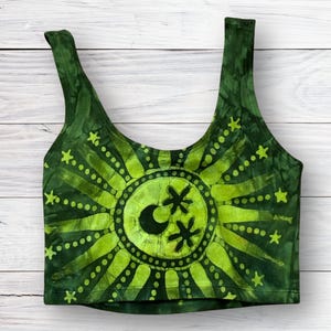 May include: A sleeveless crop top in shades of dark and lime green featuring a tie-dye effect. The center of the garment displays a large sun graphic with radiating rays, small stars, and a crescent moon with two star-like shapes in the middle.