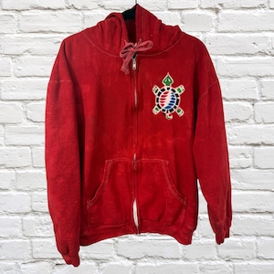 May include: A red zip-up hoodie with a drawstring hood and a kangaroo pocket. The hoodie features a colorful turtle emblem on the left chest. The emblem has a green, blue, red, and white design. The hoodie is hanging on a black hanger.