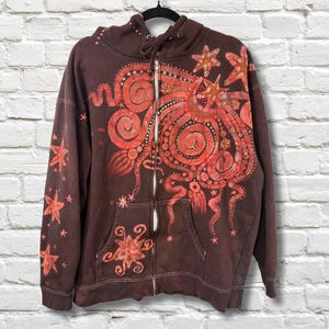 May include: A brown zip-up hoodie with a vibrant red and orange octopus design on the front and sleeves. The design includes stars and swirling patterns. The hoodie has a drawstring hood and two front pockets.