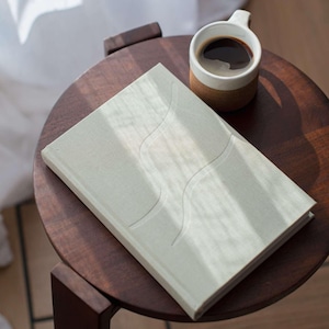 May include: A light green hardcover book with an abstract design rests on a small, dark wooden table. A white mug with a cork base and black coffee sits beside the book. The scene is lit by natural light, creating soft shadows.