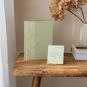 May include: A green journal with a wavy embossed design sits on a wooden table next to a card with the text "I have the resilience to help me bounce back from any setback or challenge."