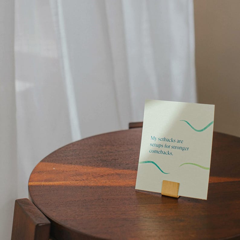 May include: A light green and white card with the text "My setbacks are setups for stronger comebacks." The card is sitting on a wooden table with a gold metal stand.