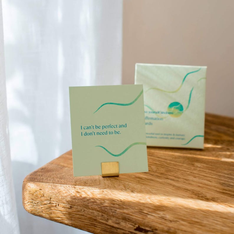 May include: A green affirmation card with the text "I can't be perfect and I don't need to be." The card is sitting on a wooden surface with a gold metal stand.
