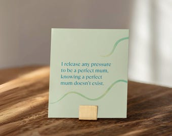 May include: A light green card with the text "I release any pressure to be a perfect mum, knowing a perfect mum doesn't exist." The card has a gold-colored base and a wavy design. The card is on a wooden surface.