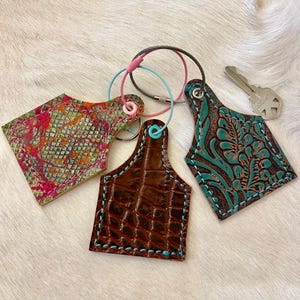 Puede incluir: Tres llaveros de cuero en varios diseños y colores. Uno es marrón con un patrón de cocodrilo, otro es turquesa con un patrón floral, y el tercero es multicolor con un diseño texturizado. Cada llavero tiene un anillo de metal y costuras coloridas.