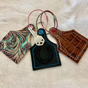 May include: Three leather keychains with metal rings. One is black with a horseshoe, blue stitching, and a key. Another is brown with a crocodile pattern and white stitching. The third is turquoise and brown with pink stitching.