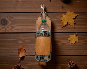 Op de afbeelding: Een lichtbruine leren waterfleshouder met een zilveren clip en een doorzichtige waterfles met een groene dop. De houder heeft blauwe stiksels en een decoratief blauw en zwart bloemmotief. De achtergrond is een bruin houten oppervlak met bladeren en dennenappels.