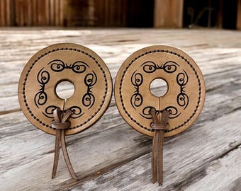 Puede incluir: Dos estribos de cuero marrón con un diseño decorativo y un borla anudada. Los estribos están sobre una superficie de madera.
