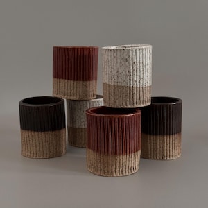 Puede incluir: Colección de seis vasos de cerámica en varios colores. Las mitades superiores son en tonos burdeos, blanco moteado con marrón y marrón oscuro. Las mitades inferiores son de color marrón claro. Cada vaso tiene detalles acanalados verticales.