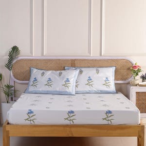 Pode incluir: Uma cama branca com uma estrutura de madeira e uma colcha branca com padrão floral. Duas fronhas combinando com um padrão floral azul estão na cama. A cabeceira é feita de cana tecida.