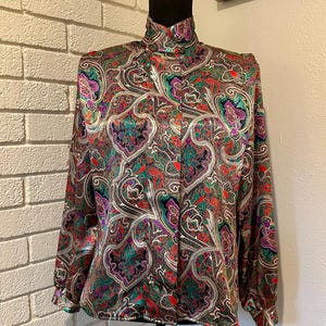 Könnte beinhalten: Eine langärmlige Bluse mit hohem Kragen und einem lebendigen Paisley-Muster in Rot-, Grün-, Lila- und Goldtönen. Die Bluse wird auf einer schwarzen Schaufensterpuppe präsentiert, die ihr aufwendiges Design und ihre seidige Textur hervorhebt. Ein Vintage-Modeartikel.