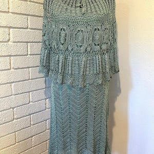 May include: A two-piece, light green crochet outfit. The top is a short-sleeved, round-neck design with a decorative pattern. The skirt is a knee-length, chevron-patterned design. The outfit is displayed on a black mannequin.