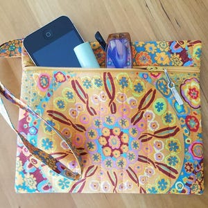 May include: A quilted fabric wristlet pouch featuring a vibrant, multi-colored floral mandala pattern in shades of orange, yellow, blue, and pink. The pouch has a gold-toned zipper closure, a matching wrist strap, and contains a smartphone and glasses.