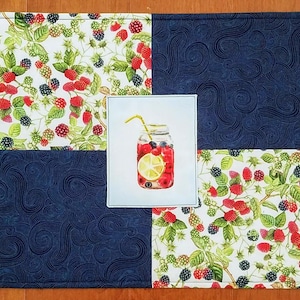 May include: A square quilted fabric mat featuring a four-panel design. Two panels display a white background with red raspberries, blackberries, and green leaves. Two panels feature a dark blue fabric with a swirling pattern. A central square shows a mason jar filled with fruit and a lemon slice.