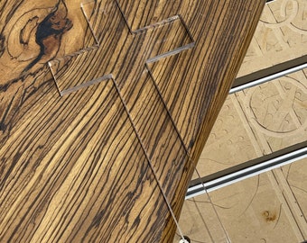 May include: A clear acrylic cross on a wooden surface with a brown and black wood grain pattern.