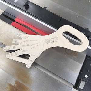 May include: A wooden hand-shaped cutout with a handle. The cutout has a detailed drawing of a hand with fingers and lines. The handle has the text "CRAFTED ELEMENTS" engraved on it.