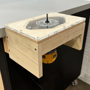 May include: A wooden router table with a clear acrylic top and a yellow and black router attached to the bottom. The router table is mounted to a workbench.