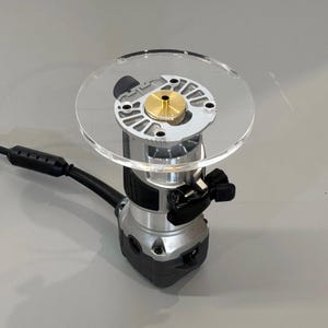 May include: A silver and black electric router with a clear plastic base and a brass-colored center. The tool has a black power cord and a black handle. The router is designed for woodworking and shaping materials.