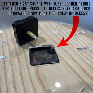 May include: A close-up of a clock movement mechanism with a black square housing and a gold-colored shaft. The mechanism is mounted on a wooden surface with a clear plastic cover. The text "S" is visible on the mechanism.