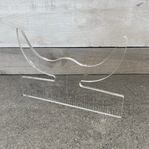 May include: A clear acrylic hat stand with a curved top and a ruler on the base. The stand is designed to hold hats in place for display or storage.