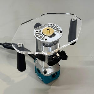 May include: A clear acrylic router table with a black router attached. The router has a brass insert and a black switch. The table has two black handles.