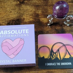 May include: A purple box with the text "Absolute Affirmations" and a pink illustration of a heart being hugged. The box is on a wooden surface next to a purple affirmation card with the text "I Embrace the Unknown" and a silhouette of a roller coaster against a sunset.