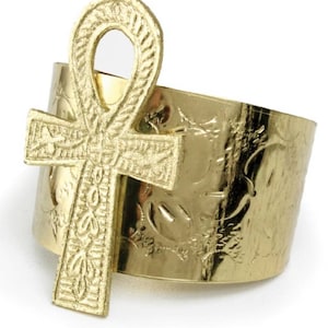 May include: A gold-toned ring with an ankh symbol design. The ring is wide and has an engraved pattern.