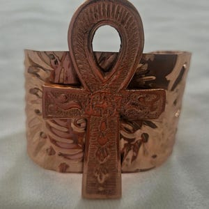 May include: A copper-colored cuff bracelet featuring an embossed Ankh symbol. The bracelet has a textured surface with floral-like patterns. The Ankh is a cross with a loop at the top, a symbol of life.
