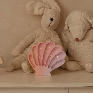 May include: A decorative seashell-shaped night light in pink and purple hues, with small golden lights. Two plush animal toys, a bunny and a lamb, sit beside the light. A natural rattan rainbow shelf hangs above.