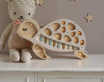 May include: A wooden turtle-shaped night light with a cream-colored shell and a light-colored wooden frame. The shell has numerous circular cutouts with small lights inside. The turtle has four legs and a smiling face. A stuffed lamb toy is next to the light.
