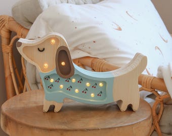 May include: A wooden dachshund-shaped night light with a light blue body and cherry accents. The dog's head and legs are natural wood, and the light has several small, warm lights. It sits on a small wooden stool.