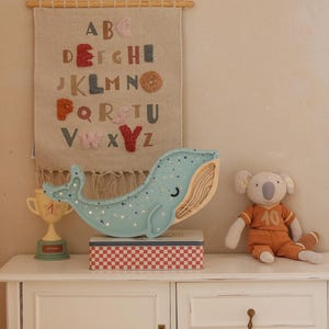 May include: A light blue whale-shaped night light with silver glitter sits atop a red and white checkered box. A koala bear doll in an orange jersey and a gold trophy are also on the white cabinet. A fabric alphabet wall hanging is behind the whale.