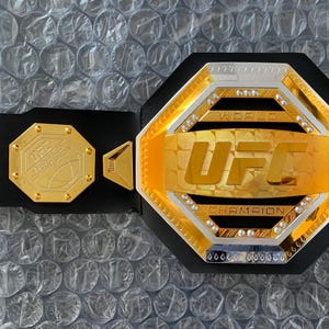 May include: A gold and silver UFC championship belt with a black strap. The belt features the UFC logo and the words "World Champion".