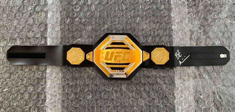 May include: A black and gold UFC championship belt with a black strap and a signature on the strap. The belt features a large octagon-shaped center plate with the word "UFC" in white letters.