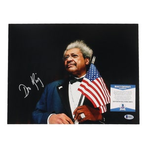 Puede incluir: Una fotografía en blanco y negro de un hombre con traje sosteniendo una bandera estadounidense. Está mirando hacia la derecha y tiene una expresión seria en su rostro. La firma del hombre, "Dr. King", está en la esquina inferior izquierda de la foto. Un Certificado de Autenticación Beckett está en la esquina inferior derecha de la foto.
