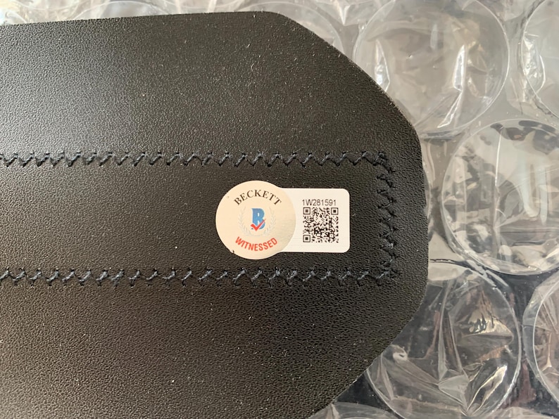 May include: A black leather item with a Beckett Witnessed sticker and a barcode sticker with the number 1W281591.