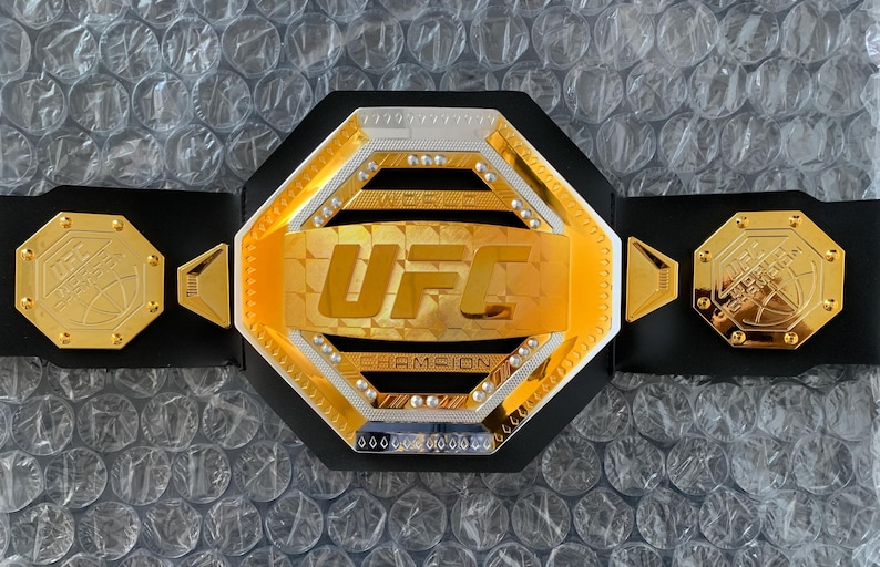 May include: A gold and silver UFC championship belt with a black strap. The belt features the UFC logo and the words "World Champion".