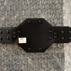 May include: Black leather wrestling championship belt with a large octagonal center plate and metal studs. The belt has a velcro closure and the text "WWE" is printed on the center plate.