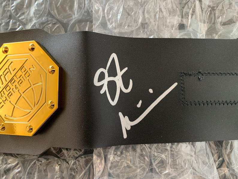 May include: A black leather championship belt with a gold octagonal plate featuring a basketball and the text "The School of Hoop Championship". The belt is signed in white ink with the name "Alex".