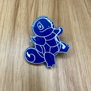May include: A blue and white enamel pin featuring a cartoon turtle with a shell and a tail. The turtle is outlined in white and has a white eye.