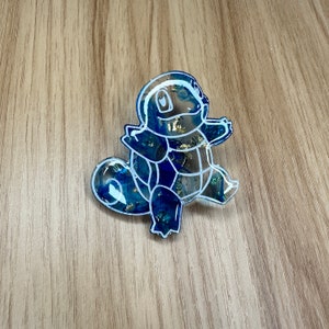 May include: A blue and white resin charm of a Pokemon character, Squirtle. The charm is outlined in white and has a blue and gold marbled effect.