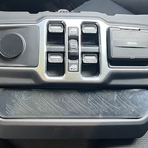May include: Black plastic car interior panel with silver trim and buttons. The panel has a round button, four square buttons, and a rectangular button labeled "MEDIA".