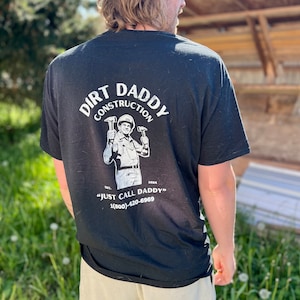 May include: Black t-shirt with a white graphic of a construction worker holding two hammers. The graphic says "Dirt Daddy Construction" and "Just Call Daddy" with a phone number.
