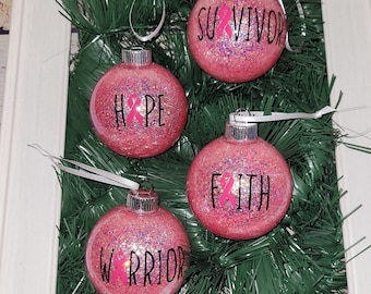 May include: Four pink glitter Christmas ornaments with silver caps and white ribbons. Each ornament has a word in black lettering and a pink ribbon: "HOPE", "SURVIVOR", "FAITH", and "WARRIOR". The ornaments are displayed on a bed of green faux pine needles.