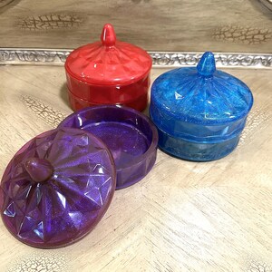 May include: Three decorative resin trinket boxes in red, blue, and purple. Each box has a faceted design and a matching lid with a small knob. The purple box has its lid open, revealing the interior.
