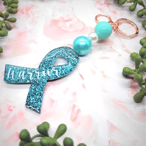 May include: A teal glitter ribbon keychain with the word "Warrior" in white script. The keychain has a beaded accent with two teal beads and a white pearl, attached to a rose gold keyring. The background is a soft pink and white floral pattern.
